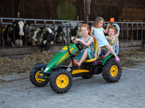 Drie kinderen op een groene traptractor bij een koeienstal in Feather Down De Kalverweide, Overijssel.