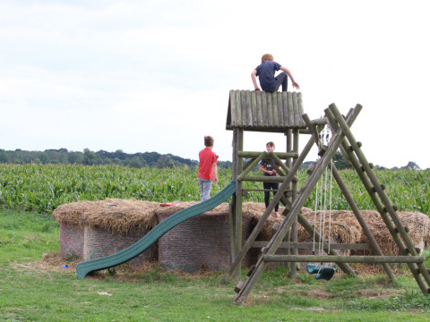 Kinder spielen auf einem Spielplatz mit Schaukel, Rutsche und Strohballen neben einem Maisfeld bei Feather Down College Farm.