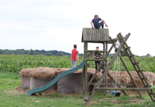 Kinder spielen auf einem Spielplatz mit Schaukel, Rutsche und Strohballen neben einem Maisfeld bei Feather Down College Farm.