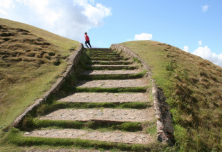 Escalier en pierre montant une colline herbeuse sous un ciel bleu, avec une personne en veste rouge en haut.