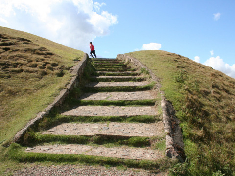 Escalier en pierre montant une colline herbeuse sous un ciel bleu, avec une personne en veste rouge en haut.
