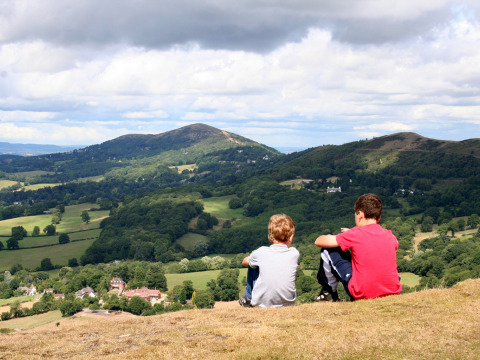 Two boys sit on a hillside overlooking the scenic green valleys at Feather Down Hollings Hill, West Midlands.