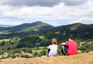 Zwei Kinder sitzen auf einem Hügel mit Blick auf die grünen Hügel bei Feather Down Hollings Hill, West Midlands.