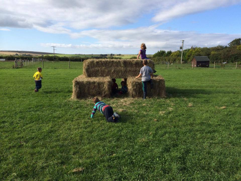 Niños juegan en pacas de heno al aire libre bajo el cielo azul en Feather Down Warren Farm, Suroeste de Inglaterra.