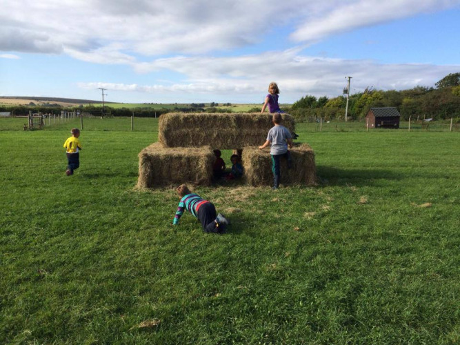 Kinderen spelen buiten op hooibalen onder een blauwe lucht bij Feather Down Warren Farm in Zuidwest-Engeland.