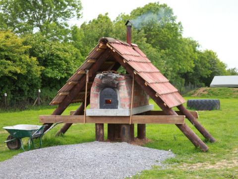 Horno de ladrillo al aire libre con techo de tejas en Feather Down Warren Farm rodeado de césped y árboles.