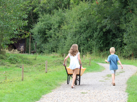 Due bambini camminano su un sentiero di ghiaia, una bambina spinge una carriola, circondati dal verde a Feather Down Upper Shadymoor Farm.