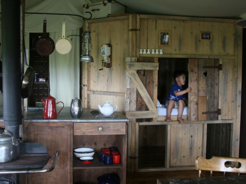 Interior de glamping con paredes de madera rústica, utensilios de cocina y niño en una cama empotrada.