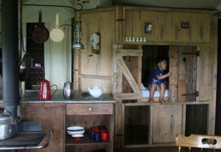 Interior de glamping con paredes de madera rústica, utensilios de cocina y niño en una cama empotrada.