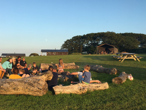 Familias y niños sentados en troncos en Feather Down Treganhoe Farm, un parque vacacional en el suroeste de Inglaterra.