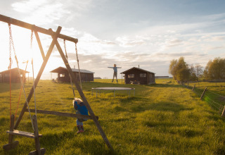Børn leger på en gynge og en trampolin på en solrig mark med hytter på Feather Down Hoeve den Overdraght.