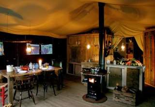 Cucina rustica in tenda glamping, con stufa a legna, tavolo in legno, lanterne e verdure fresche.