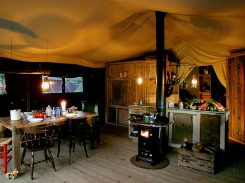 Cucina rustica in tenda glamping, con stufa a legna, tavolo in legno, lanterne e verdure fresche.