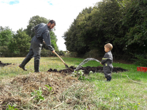 Un hombre y un niño con monos de trabajo y botas riegan y trabajan juntos en un jardín rodeado de árboles.