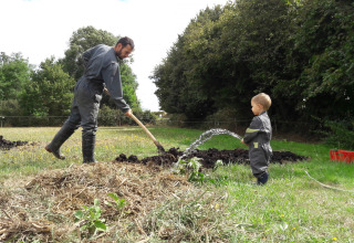 Een man en een kind in werkkledij en laarzen werken samen in de tuin, omringd door gras en bomen.