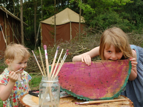 Two children play with paint brushes and a painted log slice outdoors at Feather Down Layer Marney Tower park.