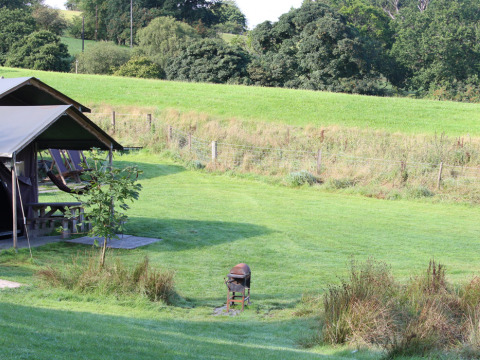 Vista di Feather Down Pant y March Farm in Galles con prati verdi, tenda glamping e barbecue esterno.