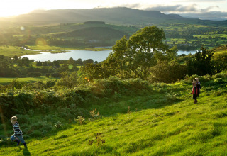 Bambini giocano su una collina erbosa con vista sul lago e sulle colline a Feather Down Pant y March Farm, Galles.