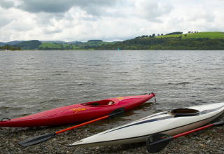 Due kayak sono sulla riva sassosa di un lago con colline verdi sullo sfondo a Feather Down Pant y March Farm, Galles.