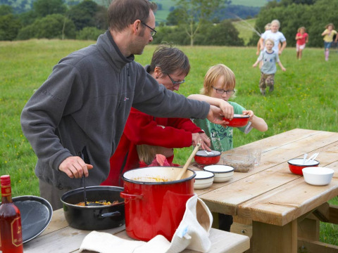 Famiglia che cucina all'aperto su un tavolo da picnic a Feather Down Pant y March Farm, Galles, Regno Unito.
