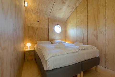 Kleine, gezellige slaapkamer met houten wanden, een tweepersoonsbed, nachtlampje en rond raam.