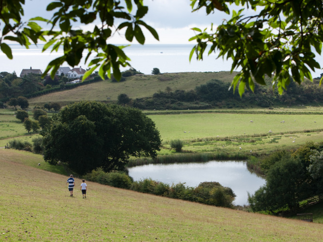 Two people walk down a hill overlooking a pond, fields, and houses at Feather Down Lunsford Farm, England.