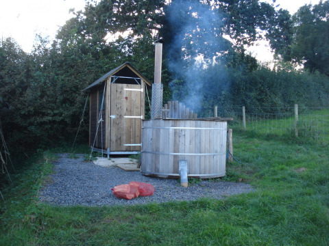 An outdoor wooden hot tub emitting smoke beside a wood shed at Feather Down Billingsmoor Farm in England.