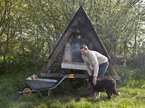 Man pets a dog beside an outdoor oven and a wheelbarrow of firewood at Feather Down Billingsmoor Farm.