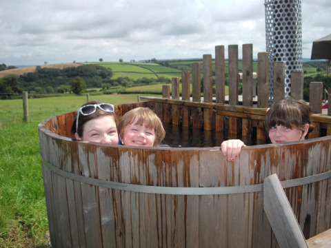 Tres niños sonríen en una bañera de madera en Feather Down Billingsmoor Farm, suroeste de Inglaterra, Reino Unido.