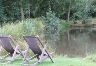 Zwei leere Liegestühle auf einer Wiese an einem Teich, umgeben von Bäumen bei BoerenBed Sunninglye Farm, England.