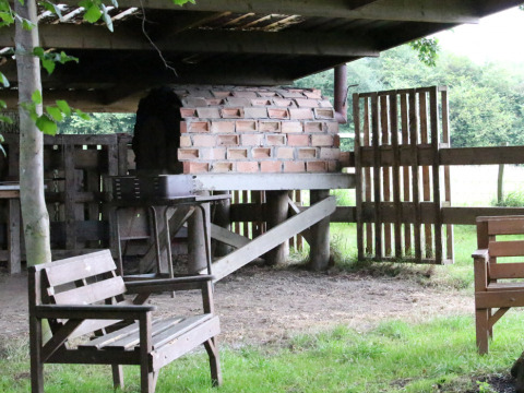 Buitenscène op BoerenBed Sunninglye Farm met een stenen oven, houten zitjes en groene natuur.