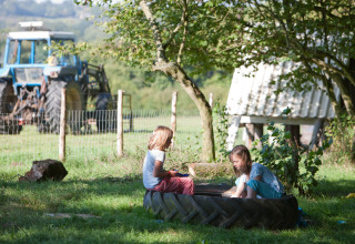 Deux enfants jouent sur l’herbe près d’un pneu de tracteur à la ferme avec tracteur en arrière-plan.