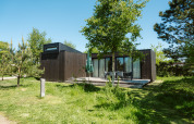 Moderna cabaña de madera oscura en Kleproos, Holiday park De Klepperstee, rodeada de césped y árboles.