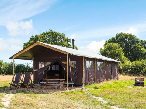 Tenda safari su un prato aperto con sedie a sdraio esterne, tavolo da picnic e alberi sullo sfondo sotto il cielo blu.