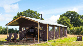 Safari tent op een open veld met ligstoelen buiten, picknicktafel en bomen op de achtergrond onder een blauwe lucht.