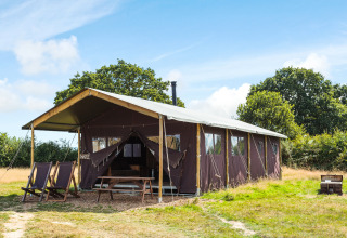 Safari tent on an open field with deck chairs outside, picnic table, surrounded by trees and blue skies.