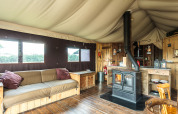 Interno di una tenda safari con pavimento in legno, stufa, divano rustico e accogliente arredamento naturale.