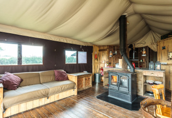 Interno di una tenda safari con pavimento in legno, stufa, divano rustico e accogliente arredamento naturale.