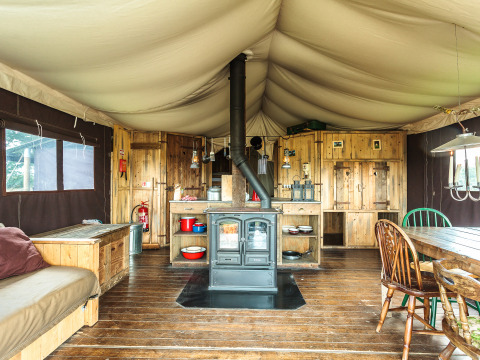 Interieur van een safaritent met houten vloer, houtkachel, rustieke keuken en grote eettafel.