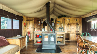 Interno di tenda safari con pavimento in legno, stufa, zona cucina rustica e tavolo da pranzo grande.