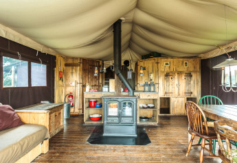 Interno di tenda safari con pavimento in legno, stufa, zona cucina rustica e tavolo da pranzo grande.