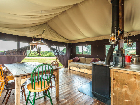 Intérieur de tente safari avec table en bois, chaises variées, poêle à bois, canapé et vue sur la nature.