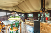 Innenraum eines Safarizelts mit Holztisch, verschiedenen Stühlen, Kaminofen, Sofa und Naturblick.