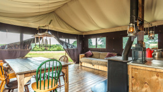 Innenraum eines Safarizelts mit Holztisch, verschiedenen Stühlen, Kaminofen, Sofa und Naturblick.