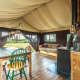 Safari tent interior with wooden table, assorted chairs, wood stove, couch, and view of nature outside.