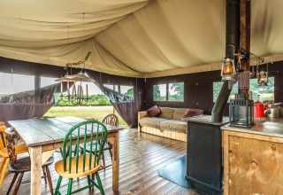 Interno di tenda safari con tavolo in legno, sedie diverse, stufa, divano e vista sulla natura esterna.