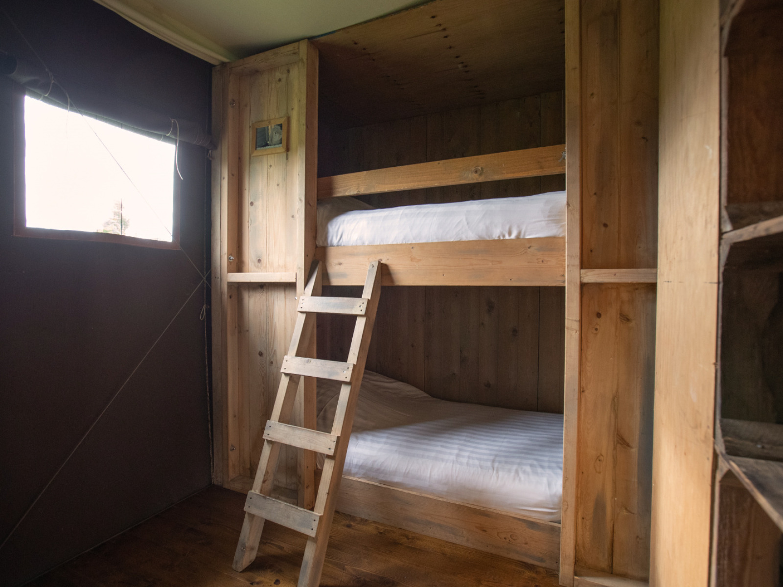 Houten stapelbedden met ladder in een safaritent, rustieke sfeer en daglicht via een raam aan de zijkant.