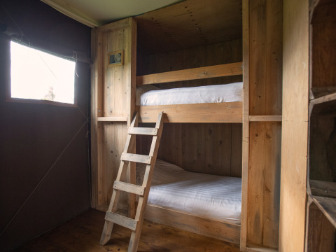 Houten stapelbedden met ladder in een safaritent, rustieke sfeer en daglicht via een raam aan de zijkant.