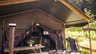 Safari-Zelt mit offener Veranda, Holztisch, Liegestühlen und Blick auf grüne Wiese und Wald.