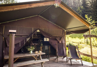 Tente safari avec terrasse ouverte, table en bois, chaises longues et vue sur la forêt verdoyante.